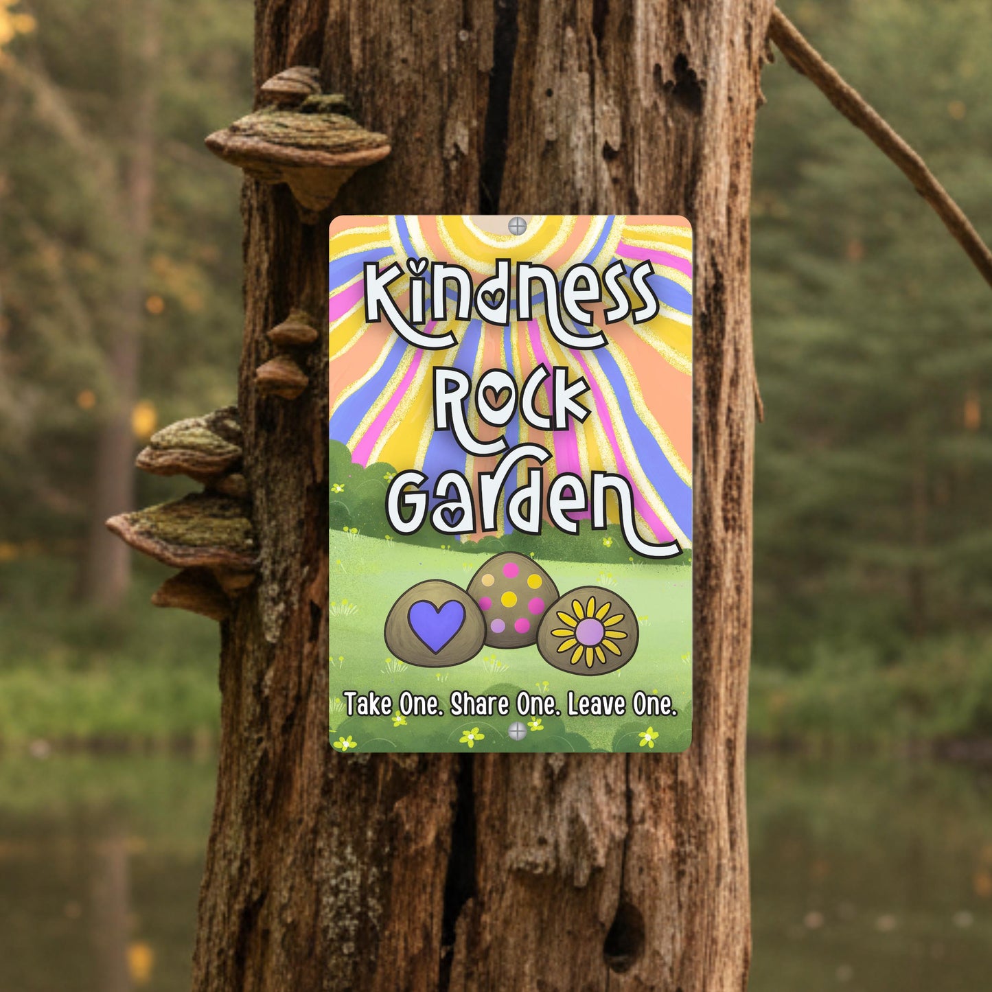 A vertical metal sign mounted on a tree, with the words "Kindness Rocks Garden" in a colorful, retro font and groovy background. The sign features three illustrated rocks with a heart, polka dots, and a sunflower. The text "Take One. Share One. Leave One." is at the bottom.